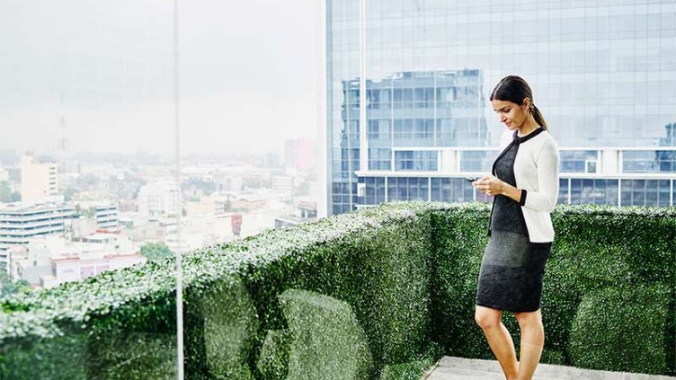 Smiling female business executive standing on office terrace looking at information on smartphone